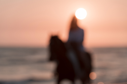 Blurred photo. A woman in summer clothes enjoys riding a horse on a beautiful sandy beach at sunset. Selective focus. High-quality photo