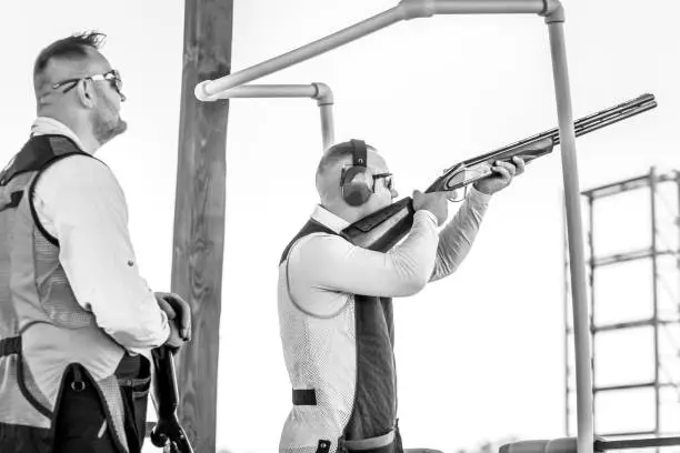 Photo of Two adult mans in sunglasses, protective headphones and a rifle vest practicing fire weapon shooting.