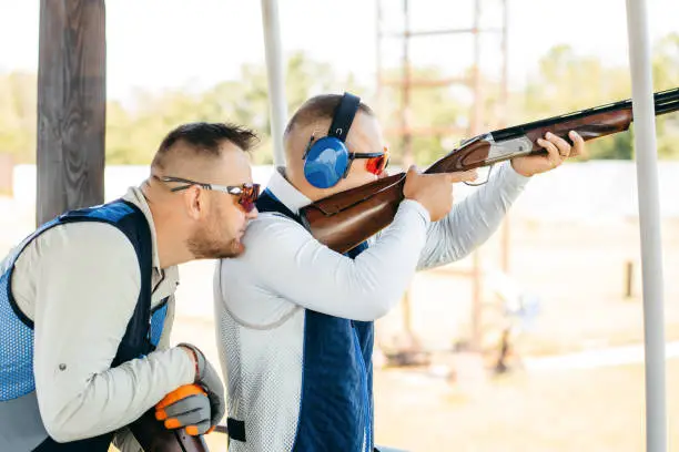 Photo of Two adult mans in sunglasses, protective headphones and a rifle vest practicing fire weapon shooting.