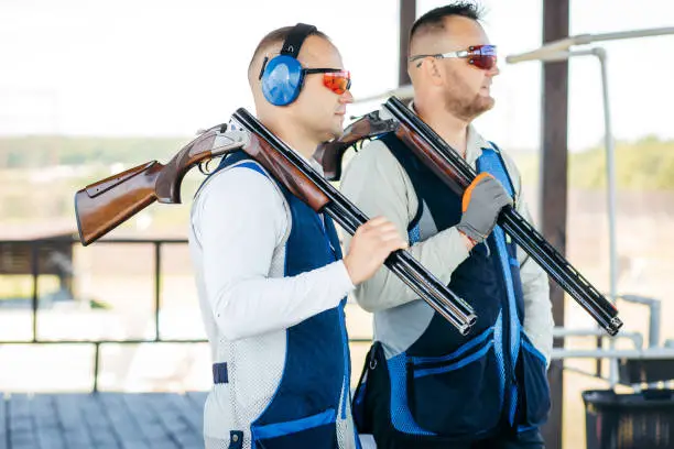 Photo of Portraits of two adult mans in sunglasses, protective headphones and a rifle vest practicing fire weapon shooting.