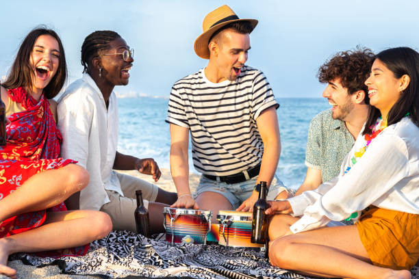 grupa wielorasowych przyjaciół bawiących się na plaży, śmiejących się na głos, grających w bongo i pijących razem piwo. - party beach indian ethnicity adult zdjęcia i obrazy z banku zdjęć