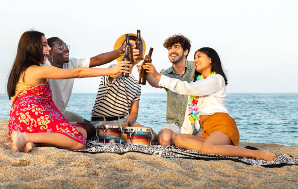 szczęśliwa, zróżnicowana grupa przyjaciół wznosząca toast przy piwie razem na plaży. wielorasowi młodzi ludzie bawiący się na świeżym powietrzu. - party beach indian ethnicity adult zdjęcia i obrazy z banku zdjęć