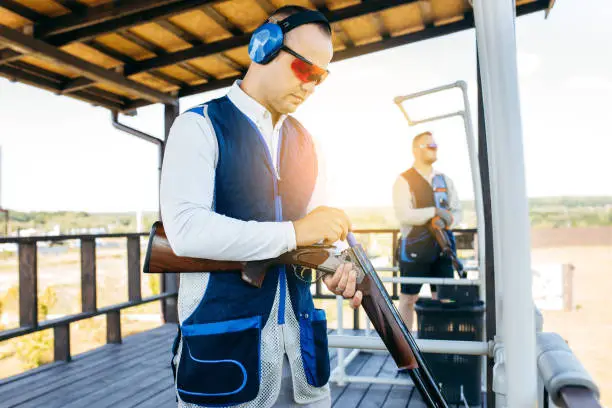 Photo of Two adult mans in sunglasses, protective headphones and a rifle vest practicing fire weapon shooting.