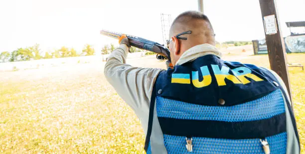 Photo of Adult man in sunglasses and a rifle vest practicing fire weapon shooting. Young experienced male aiming shotgun in outdoor.