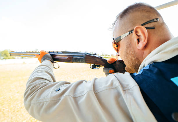 istock Adult man in sunglasses and a rifle vest practicing fire weapon shooting. Young experienced male aiming shotgun in outdoor. 1406891908