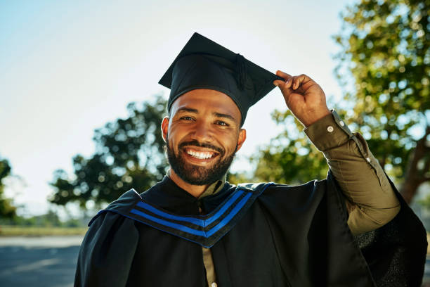 portrait of smiling graduate in gown and hat standing alone on university campus at graduation ceremony. excited, happy qualified postgrad graduating college for academic bachelor, masters and degree - grad portrait imagens e fotografias de stock