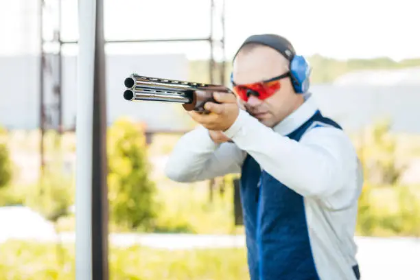 Photo of Blurred background with focus on front of shotgun barrel of adult man in sunglasses, protective headphones and a rifle vest practicing fire weapon shooting. Young experienced male aiming at the camera