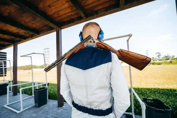 Photo of Portrait of adult man from back in sunglasses, protective headphones, rifle vest and with a shotgun on back practicing fire weapon shooting.