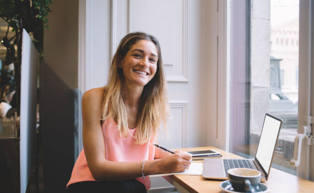 istock Positive young female author taking notes in planner and drinking coffee in cafeteria 1406896713