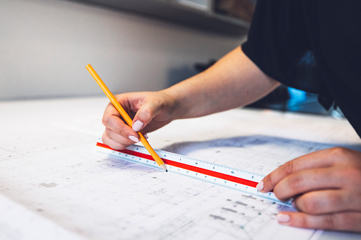 Female architect hands drawing straight line on a blueprint, using a ruler and pencil.