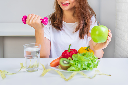 Women hands holding green apple while holing dumbbell for exercise with fruits, vegetables, water and tape measure on white table background. Selection of healthy food and exercise concept.