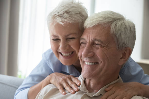 Closeup shot affectionate smiling aged wife embrace husband from behind Close up shot affectionate smiling middle aged wife embrace husband pensioner from behind massage shoulders of beloved man express love care devotion. Romantic cheerful old age couple hug touch heads dentures stock pictures, royalty-free photos & images