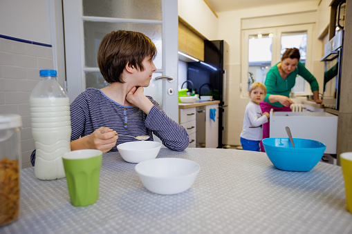 Pre-adolescent boy sitting at the dining table at home and eating his breakfast cereal with milk, looking at his brother and mother in the kitchen, family at home in the morning