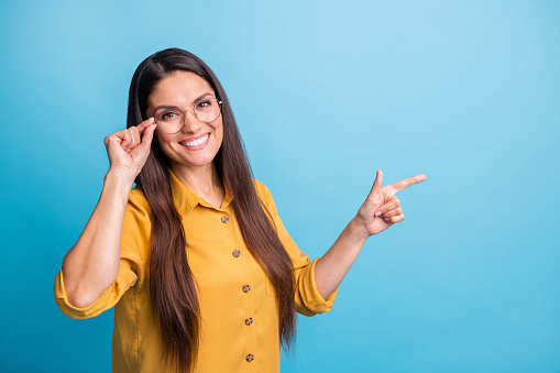 Portrait of pretty cheerful lady hand touch glasses finger indicate empty space isolated on blue color background
