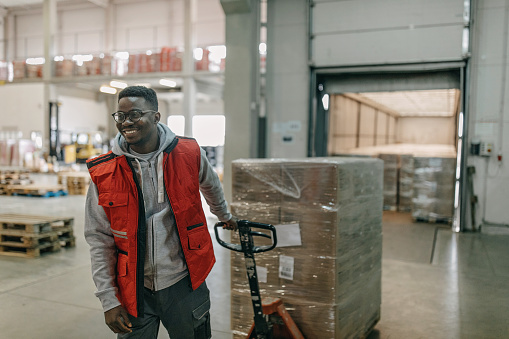 A young African American male warehouse worker is pulling a transportation cart carrying boxes, with a big smile on his face.