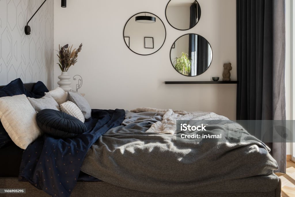 Cozy and stylish bed in modern bedroom Cozy and stylish bed with nice bedclothes in modern bedroom with decorations, round mirrors and dark window curtains Apartment Stock Photo
