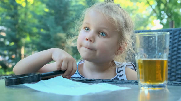 cute little girl sits thoughtfully at the table with a lens in her hand. close-up of blonde girl holding a magnifying glass in her hand and thinking about something while sitting in street cafe on the park. - magnifying glass lens holding europe imagens e fotografias de stock