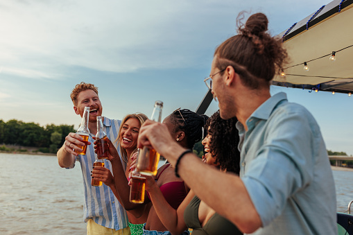 A stylish casual group of friends are at the river on a boat having beers and a good time together toasting with beers