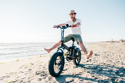 Young man on bicycle having fun with electric bike - carefree boy having fun and smiling on bicycle on the beach on a sunny day - freedom concept
