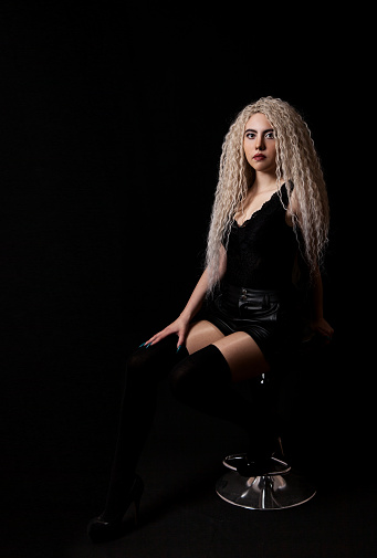 young blonde girl with long curly hair posing while sitting on a chair in the studio with black background