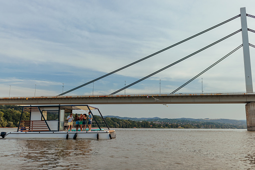 A small party  boat with a couple of people on it is floating on the Danube river next to a bridge,  while they're enjoying their leisure time.