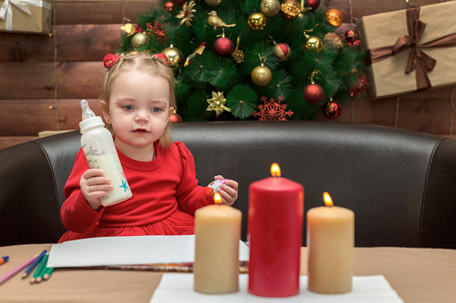 Little girl with a bottle of milk on the background of Christmas tree.