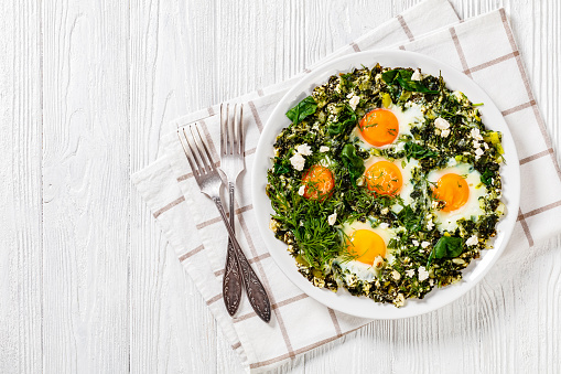 green shakshuka of spinach, leeks, green onion, crumbled feta cheese and fried eggs on white plate on wooden table with silver forks, horizontal view from above, flat lay, free space