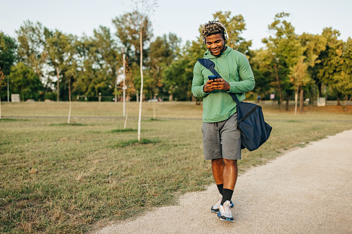 A young African American man is walking home from his practise, wearing headphones and using his phone.