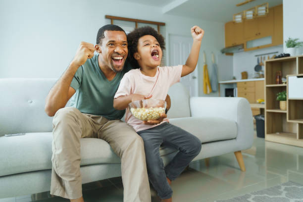 African Father and boy cheering sports games Cheerful African American Father and his little boy watching and cheering sports games competition together at home, celebrate excited football fan cheering for his team on tv stock pictures, royalty-free photos & images