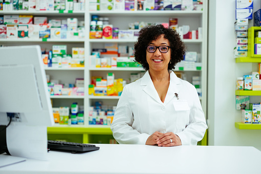 A mature pharmacist is standing at the counter smiling for the camera