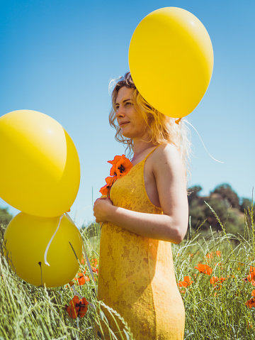 vertical portrait of beautiful woman wearing yellow dress, holding a orange poppy and some yellow balloons in a summer day