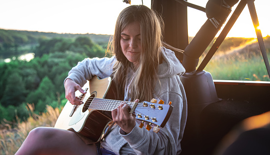 A young woman plays an acoustic guitar in a car trunk outdoors in the countryside at sunset.