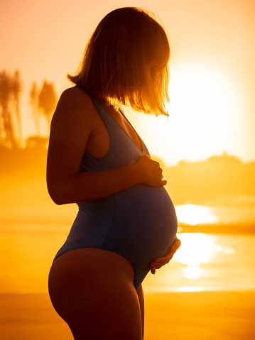Portrait of pregnant woman in swimwear posing at ocean coastline with bright sunrise or sunset