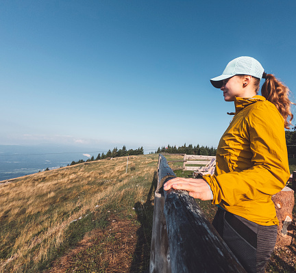 Side view of woman hiker wearing a yellow jacket and a hat, holding her hands on the fence and looking at the view.