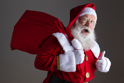 Photo of real Santa Claus with a lot of gifts on a gray background. Merry Christmas and Happy New Year! Copy space.