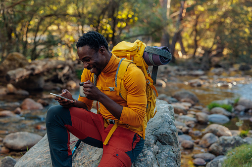 A young hiker is taking a break from hiking and reading text messages on his smartphone and drinking water.