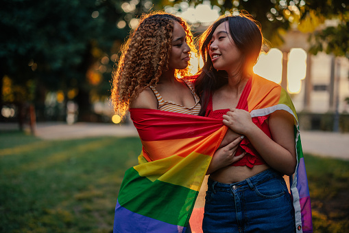 A lesbian couple with a pride flag is standing outdoor looking at each other in the park.