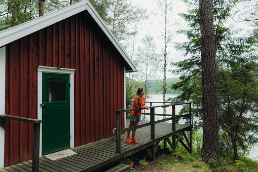 Young woman in orange acontemplating the beautiful Swedish nature, walking at the camping cabin terrace and looking at the forest and the lake