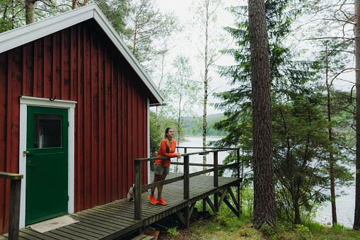 Young woman in orange acontemplating the beautiful Swedish nature, walking at the camping cabin terrace and looking at the forest and the lake