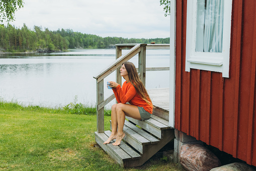 Young woman in red with long hair, sitting on the patio of the old wooden camping cabin drinking coffee, enjoying a view of the lake and the green pine woodland