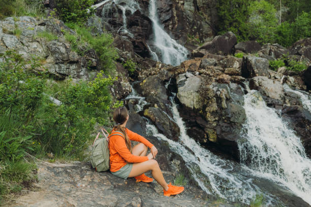 Female with backpack contemplating a view of the beautiful waterfall in Sweden Young woman backpacker in orange witting with the viewpoint of the big waterfall in the pine forest - contemplating the scenic Swedish nature during summertime norrbotten province stock pictures, royalty-free photos & images