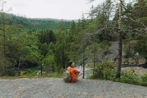 Young woman in orange with a backpack walking at the cliff enjoying a view of the scenic waterfall hidden in the green pine forest in Sweden