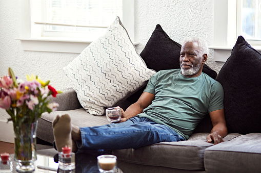 Bearded man in jeans and t-shirt sitting on sofa with feet up and looking away from camera with contented expression.