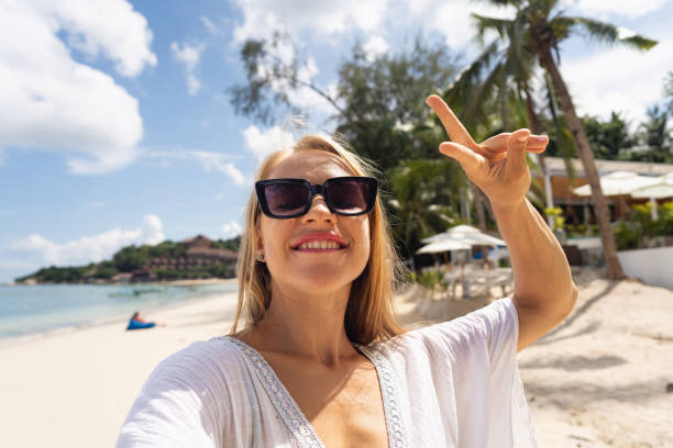 istock Young woman gesturing peace sign, while taking selfie on the beach 1406942067