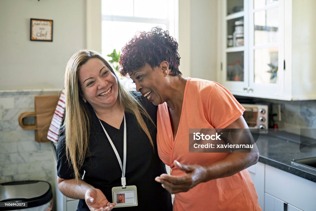 Miami senior woman and home caregiver together in kitchen Waist-up view of Black client and healthcare worker standing with arms around each other and laughing during routine house call. Community Outreach Stock Photo
