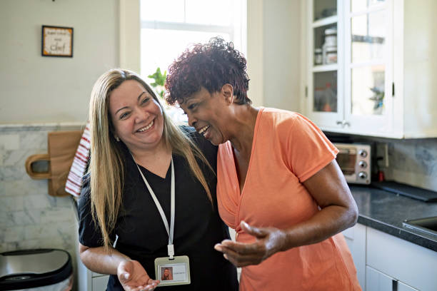 Miami senior woman and home caregiver together in kitchen Waist-up view of Black client and healthcare worker standing with arms around each other and laughing during routine house call. healthcare worker stock pictures, royalty-free photos & images