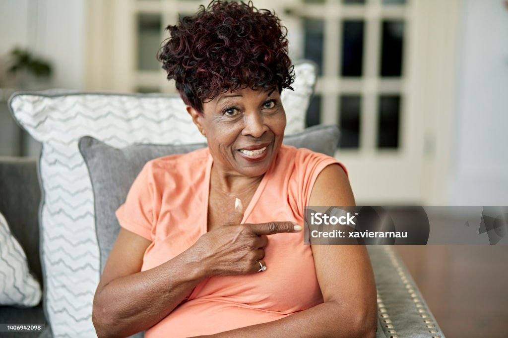 Black woman in late 70s pointing to vaccination Waist-up view of casually dressed woman sitting in family home and smiling at camera as she gestures with pride at adhesive bandage. Senior Adult Stock Photo