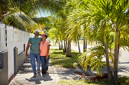 Full length view of Black couple in their 70s wearing casual clothing and approaching camera with arms around each other, talking, and laughing.