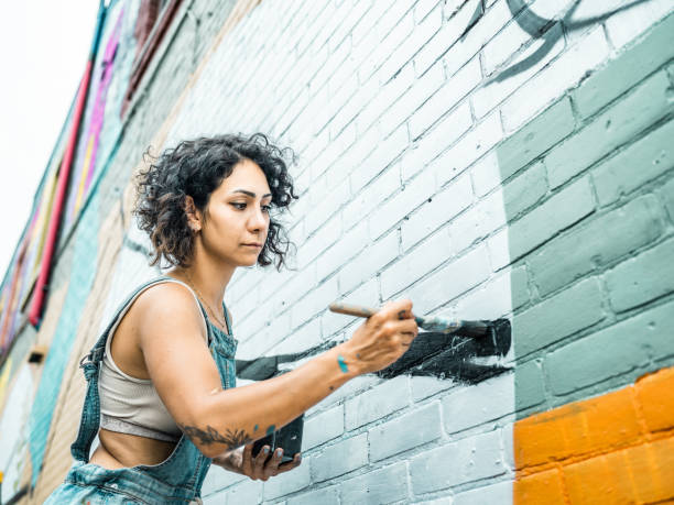 Female artist painting large wall mural Female artist painting large wall mural. She is dressed in casual work clothes. Exterior of public  downtown of large North American City. Captured from the air via drone. mural stock pictures, royalty-free photos & images