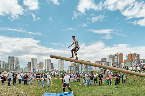 Yekaterinburg, Russia - June 25, 2022: Folk fun-competition Walking on inclined log at Sabantuy folk festival. Young Asian man coming to top of log. Spectators in clearing watching show with interest